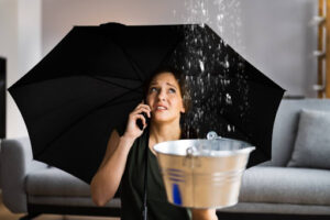 A woman using an umbrella inside a home as she uses a phone and holds a bucket up to a leak coming from the ceiling.