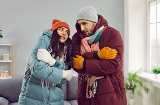 A man and a woman wearing winter gear and shivering in a home.